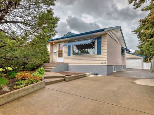 Single-story detached home with beige siding, blue shutters, front steps, landscaped yard, and long driveway leading to garage.