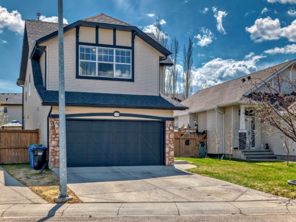 Two-story suburban house with beige siding, dark double garage door, driveway, fenced yard, and clear blue sky overhead.
