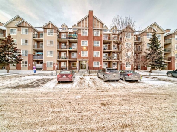 Four-story residential apartment building with balconies, brick and beige siding exterior, and snow-covered parking lot with parked cars in front.