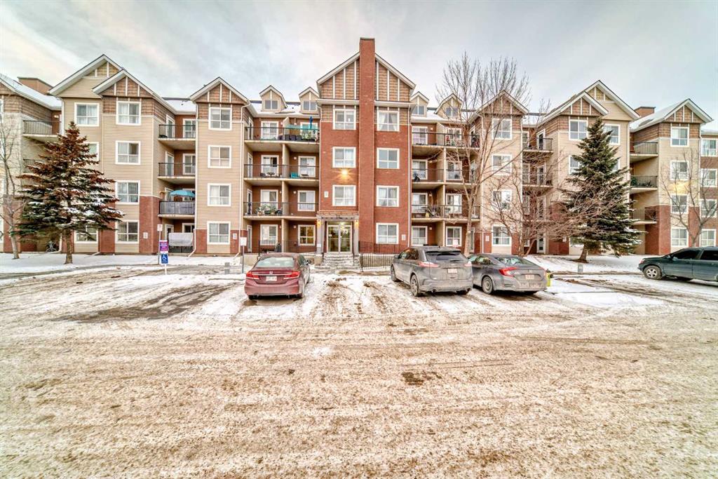 Four-story residential apartment building with balconies, brick and beige siding exterior, and snow-covered parking lot with parked cars in front.
