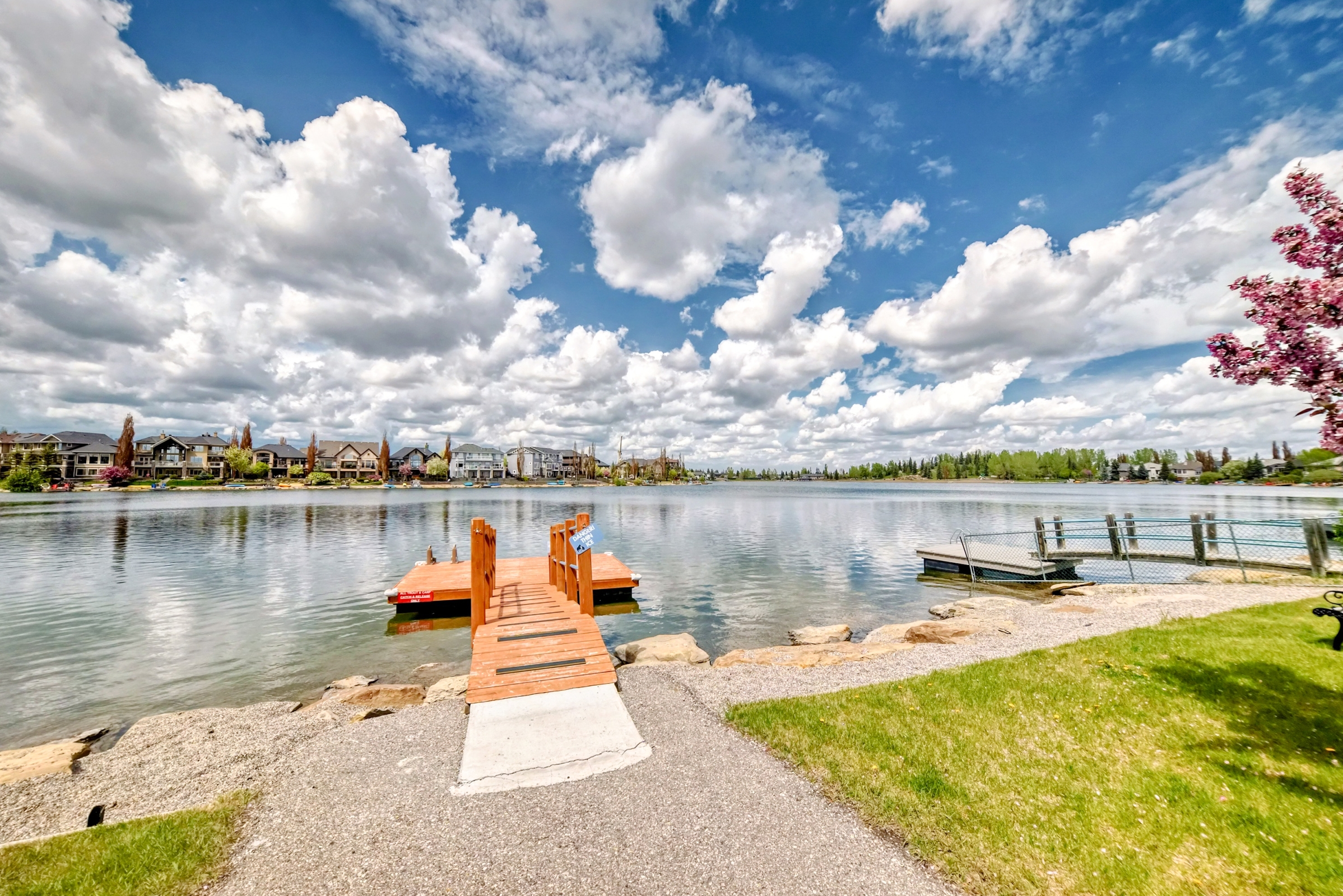 Scenic lake view with wooden dock, waterfront homes, green lawn, and bright blue sky with scattered clouds.