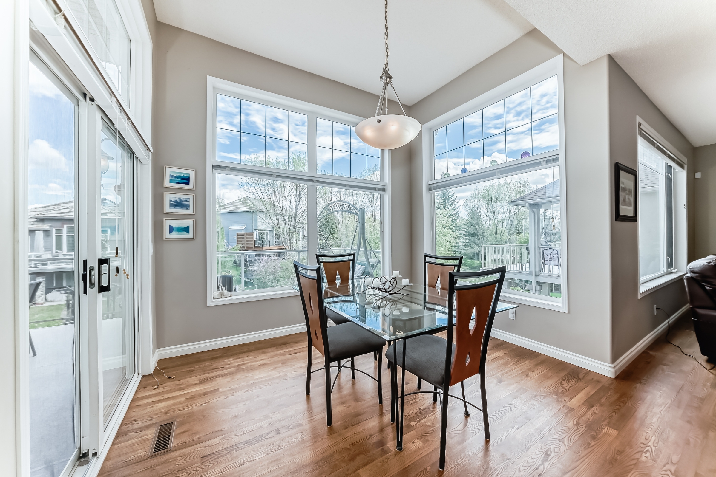 Bright dining area with glass-top table, four chairs, hardwood floors, and large corner windows overlooking backyard.